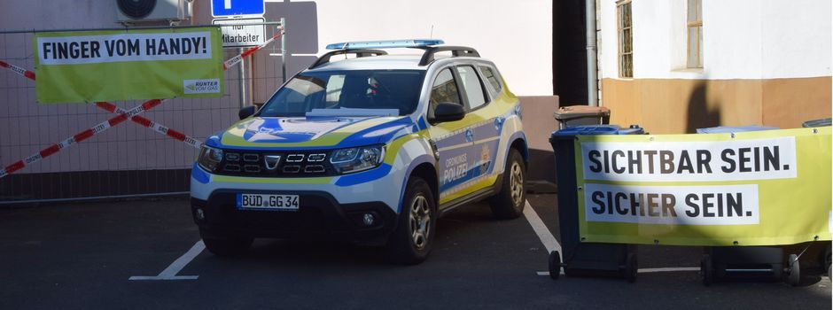 Das Bild zeigt das KfZ des Ordnungsamtes auf dem Parkplatz. Links und rechts hängen Banner mit Ordnungsamtslogos "Sichtbar sein. Sicher sein." und "Finger vom Handy!"