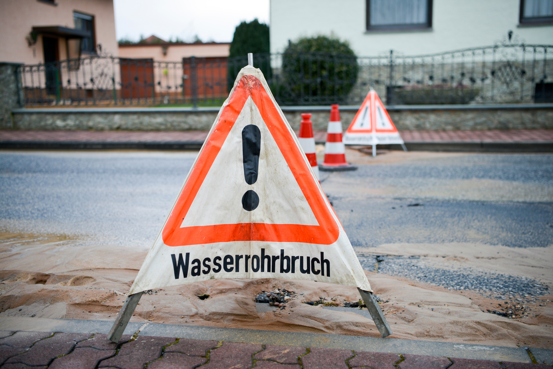 Man sieht ein Warnschild mit Aufschrift Wasserrohrbruch auf einer Straße vor einem Loch stehen.
